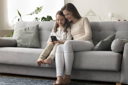 Glückliches Töchterchen, das neben seiner Mutter sitzt, Handy in der Hand, Online-Lernapp nutzt, Videospiel spielt, auf den Bildschirm schaut, lächelt, lacht, sich auf dem heimischen Sofa ausruht