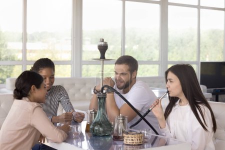 Diverse group of young millennial girls and guy enjoying leisure in hookah lounge, social communication, friendship, smoking, drinking tea, talking, speaking, relaxing on couches at table