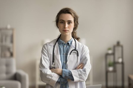 Photo for Serious confident pretty young doctor woman in white uniform coat posing with arms folded, looking at camera. Female practitioner, medical worker professional head shot portrait - Royalty Free Image