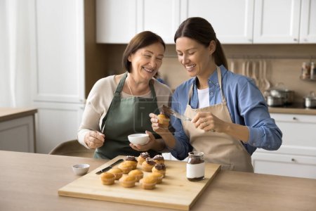 Téléchargez les photos : Joyeux maman aînée et fille adulte femme dans des tabliers profitant de l'activité de cuisson dans la cuisine à la maison, décoration de muffins, cupcakes avec chapeau crémeux au chocolat, préparation de dessert maison pour la fête - en image libre de droit