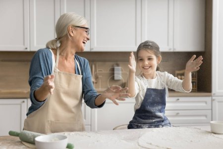 Excited grandmother and happy little granddaughter girl in aprons throwing flour over table with ingredients laughing, having fun, clapping floury hands for making cloud, playing while cooking