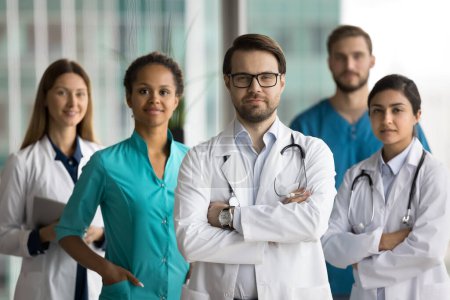 Téléchargez les photos : Médecin en chef confiant portant un manteau blanc et des lunettes debout devant une équipe diversifiée de collègues médicaux, les bras croisés, regardant la caméra. Professionnels multiethniques posant pour le portrait - en image libre de droit
