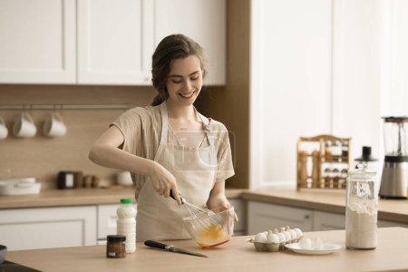 Happy beautiful young chef woman in apron preparing homemade omelet, pancakes, beating eggs in bowl, smiling, enjoying cooking, domestic culinary hobby in home kitchen