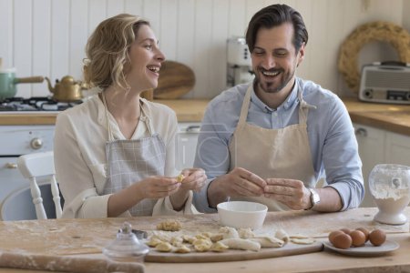 Cheerful couple in love in aprons baking in home kitchen together, enjoying cooking, culinary hobby, leisure, shaping dumplings at table with flour, dough, eggs, talking, laughing
