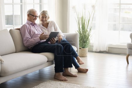 Photo for Positive elderly couple enjoying internet communication at home, holding tablet computer, looking, pointing at gadget screen, smiling, laughing, using application, online service - Royalty Free Image