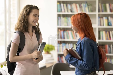 Photo for Two happy college mate girls meeting in campus library, standing together, talking, laughing, discussing class project, homework tasks, education. Cheerful students with backpacks chatting, having fun - Royalty Free Image