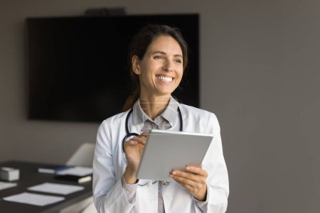 Foto de Mujer doctora adulta positiva usando tableta en la sala de conferencias de la oficina, mirando hacia otro lado con sonrisa dentada, disfrutando de la comunicación en línea, dando consulta en Internet - Imagen libre de derechos