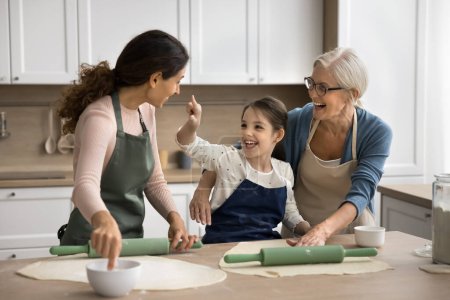 Playful cute kid girl, happy mom and granny enjoying baking, having fun, touching faces with floury finger, playing with ingredients, laughing, cooking dessert, pastry, bakery food for festive dinner
