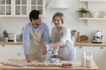 Happy positive young couple in love wearing aprons, enjoying bakery food preparation, baking pies in home kitchen, sifting flour on dough at table with ingredients, talking, smiling