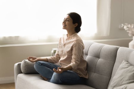 Téléchargez les photos : Joyeux paisible jeune femme indienne yogi méditant sur le canapé de la maison, gardant la pose de yoga lotus, zen mudra, geste calme de la main, souriant les yeux fermés, prenant une profonde respiration d'air frais - en image libre de droit
