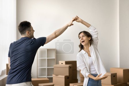Photo for Cheerful young boyfriend and girlfriend beginning living together, having fun at relocation party, dancing to music with paper boxes in background, smiling, laughing - Royalty Free Image