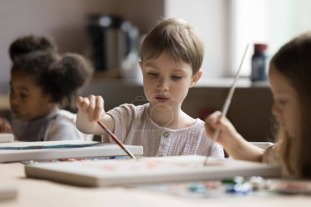 Photo for Focused cute little kid drawing on class in group of classmates, mixing colors on palette with paintbrush, sitting at table with canvas, oil paints, training artistic skills in art school studio - Royalty Free Image