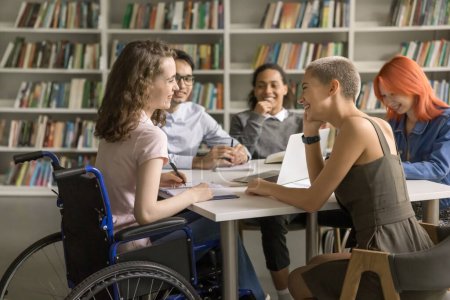 Téléchargez les photos : Jeune étudiante handicapée coopérant à un projet de classe à la bibliothèque avec des amis du collège, en fauteuil roulant, assise à table, parlant, bavardant avec des camarades de classe, riant - en image libre de droit