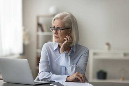 Aged businesswoman sit at workplace desk look into distance, distracted from work, feel concerned about upcoming deadline, task, project, ponder regarding career path, retirement plans or future goals