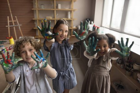 Photo for Cheerful cute multiethnic kids posing at camera in creative craft studio, showing painted palms, smiling, laughing, having fun. Pupils enjoying leisure time on artistic class - Royalty Free Image