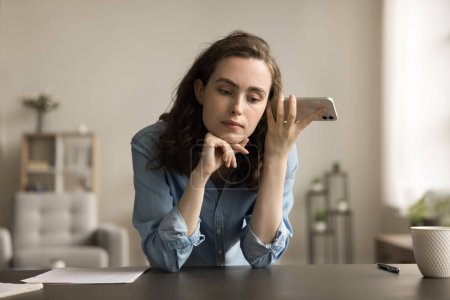 Serious freelancer employee girl holding mobile phone at head, listening to sound, voice message, audio file, leaning hands on home workplace table, thinking, touching chin