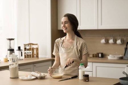 Happy dreamy pretty young woman cooking homemade breakfast in kitchen, beating fresh milk and eggs for omelet in bowl, looking away, smiling, laughing, enjoying culinary activities