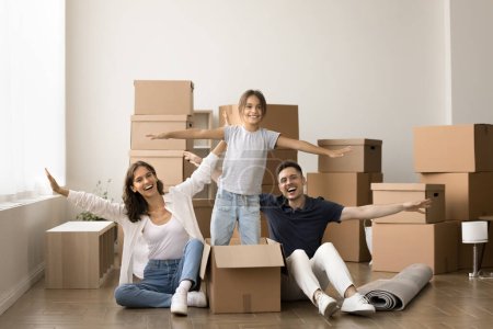 Photo for Happy little kid girl dancing in cardboard box, standing in paper container with open hands near cheerful mom and dad sitting on floor. Young couple and child enjoying moving activity - Royalty Free Image