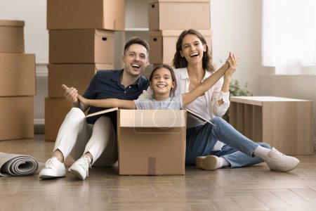 Photo for Happy mom and dad playing with joyful kid girl sticking out from moving box, sitting on floor with relocation packages behind, looking at camera, smiling, laughing, enjoying being in new home - Royalty Free Image
