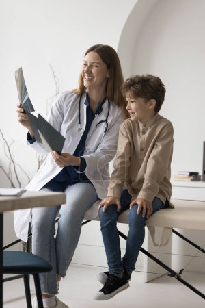 Photo for Cheerful practitioner reviewing Xray scan, shot of bones of little patient boy, feeling happy, smiling, laughing. Kid meeting with pediatrician doctor after radiography screening examination - Royalty Free Image