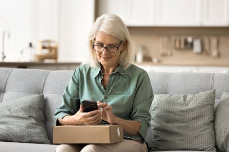 Positiv zufrieden reife Internet-Shop-Kundin im Ruhestand nutzt Handy über Karton mit Waren aus dem Geschäft, gibt positives Feedback, tippt auf Online-Bewerbung
