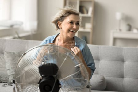 Téléchargez les photos : Femme âgée insouciante et joyeuse profitant de la fraîcheur du ventilateur, du ventilateur, du refroidisseur, assise à l'air frais soufflant du ventilateur, utilisant un appareil ménager, un équipement de refroidissement - en image libre de droit