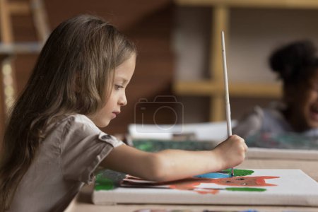 Photo for Focused cute pretty art school pupil girl drawing cartoon animal on canvas, studying creativity on painting class, holding paintbrush, sitting at big table in classroom. Side view - Royalty Free Image