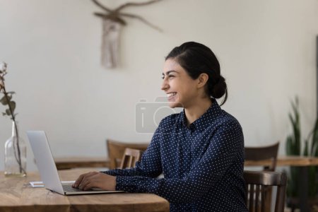 Foto de Alegre millennial independiente india mujer de negocios que trabaja en el ordenador portátil en la oficina en el hogar, sonriendo, mirando hacia otro lado, sentado en la mesa de trabajo, pensando en el proyecto exitoso visión futura, idea creativa - Imagen libre de derechos