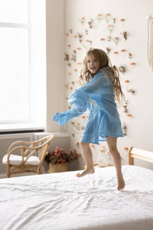 Cheerful active sweet child girl in oversized shirt dancing to music in bedroom, jumping on bed with flying cloth, hair. Happy kid enjoying motion, moving, playing at home, having fun