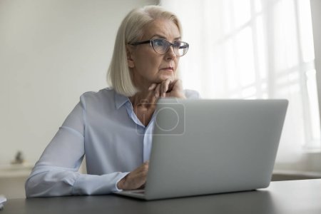 Photo for Pensive older businesswoman sit at desk, working on laptop, think over marketing report at workplace, feel doubt, analyze agreement terms, make decision, ponder task or strategy. Brainwork, technology - Royalty Free Image