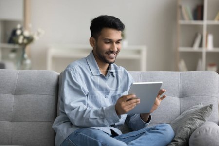 Photo for Positive attractive young Indian entrepreneur man using Internet technology, smart home application, resting on cozy sofa, holding digital tablet, reading electronic book on display, enjoying leisure - Royalty Free Image