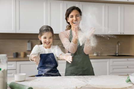 Excited mom and cheerful little daughter girl in aprons throwing flour while baking in home kitchen, having fun, laughing, clapping floury hands, enjoying culinary hobby, childhood, motherhood