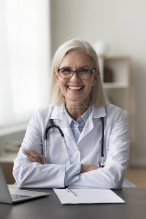 Photo for Vertical portrait of smiling mature physician or cardiologist posing for camera seated at workplace desk with laptop. Career growth, professional medical services, workflow with usage of modern tech - Royalty Free Image