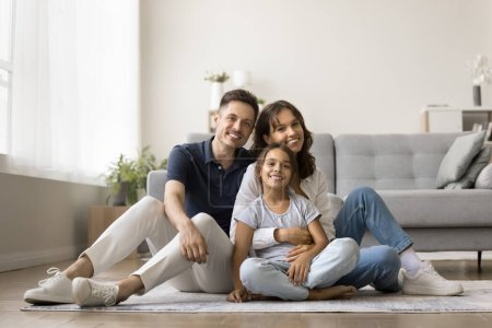 Photo for Cheerful family couple holding beautiful tween daughter girl in arms, sitting on heating floor in modern home interior, looking at camera for portrait, smiling, enjoying leisure in new apartment - Royalty Free Image