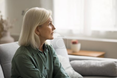 Téléchargez les photos : Femme âgée blonde frustrée solitaire regardant loin avec un regard inutile, assise sur le canapé à la maison seule, souffrant de dépression, d'apathie, de désespoir, se sentant triste, fatiguée, ennuyée - en image libre de droit