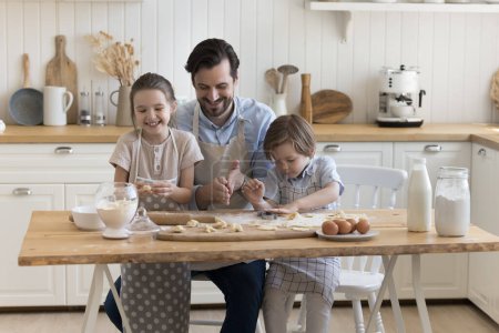 Photo for Cheerful dad teaching positive little children to bake, shaping dumplings over floury table, with dough, eggs, milk, smiling, laughing, enjoying culinary hobby, family leisure - Royalty Free Image
