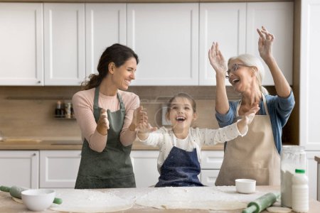 Foto de Alegre niña emocionada, mamá y la abuela lanzando harina en polvo sobre la mesa de hornear, aplaudiendo las manos, haciendo nube harinosa, riendo, divirtiéndose, disfrutando del ocio familiar, tiempo de juego, pasatiempo de cocina - Imagen libre de derechos