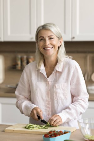 Foto de Mujer jubilada bastante madura alegre preparando ensalada para la cena en la cocina casera, mirando a la cámara con sonrisa dentada, cortando pepino, rebanando verduras frescas en la tabla de cortar - Imagen libre de derechos