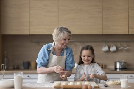 Photo for Positive granny and granddaughter kid in aprons preparing pies, cookies, biscuits in kitchen, shaping, kneading dough on floury table and board. Grandma teaching girl to bake - Royalty Free Image