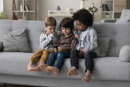 Tres diversos niños de la generación Z usando el teléfono móvil de la aplicación de aprendizaje, sentado en el sofá en casa solo. Hermanos pequeños, chicos haciendo llamadas, jugando videojuegos, viendo contenido en línea, navegando por Internet