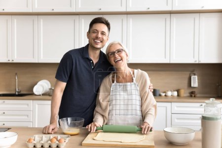 Happy younger man and elder woman cooking homemade bakery food, rolling raw dough for pitta, looking at camera, hugging, smiling, laughing. Happy senior mother and adult son domestic family portrait
