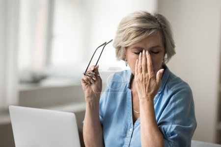 Photo for Tired blonde elderly woman taking glasses off at workplace with computer, feeling overworked, touching eyelids, face with closed eyes, suffering from headache, weak eyesight, bad vision - Royalty Free Image