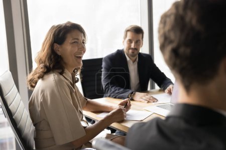 Photo for Positive mature employee woman discussing ideas for successful project, brainstorming with colleagues and male boss, sitting at meeting table, laughing, enjoying teamwork, cooperation - Royalty Free Image