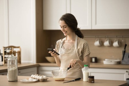 Happy beautiful young chef woman using smartphone over table with bakery food ingredients, preparing breakfast, taking picture, photo, cooking pancakes, omelet, reading online recipe on Internet