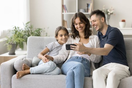 Photo for Happy young parents and cute daughter kid watching content on smartphone, using gadget for online communication, taking family selfie on cozy home couch, enjoying Internet technology - Royalty Free Image