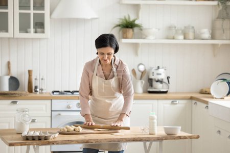 Photo for Retired mature woman wear apron cooking alone standing in modern cozy kitchen, prepare dough for homemade pastry, enjoy cookery process at home. Chores, housework, family recipe preparation, culinary - Royalty Free Image
