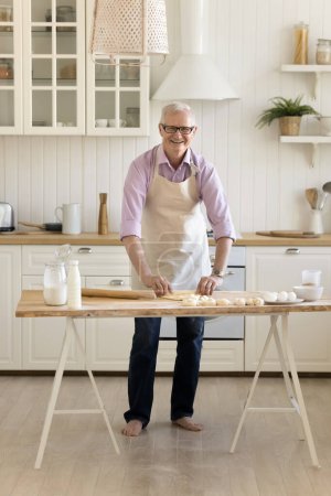 Photo for Cheerful mature grandpa engaged in culinary activity baking homemade dessert at home, standing at table with flour, milk, eggs, rolling dough, looking at camera, smiling. Vertical shot portrait - Royalty Free Image