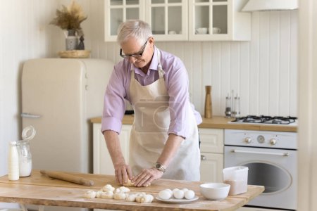 Senior baker man wearing domestic apron patting raw dough on floury table, preparing fresh buns, enjoying culinary hobby, cooking pastry dessert in home kitchen