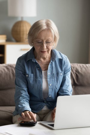 Téléchargez les photos : Focalisé vieille femme retraitée ordinateur portable payer des factures en ligne, vérifier les soldes financiers, le calcul du budget mensuel, les frais, à l'aide d'un ordinateur, calculatrice à la maison, assis sur le canapé. Plan vertical - en image libre de droit