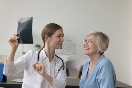 Photo for Happy doctor woman showing Xray scan to elder patient, explaining shot of bones, radiography screening film to senior woman in hospital office. Old lady visiting doctor for medical checkup - Royalty Free Image